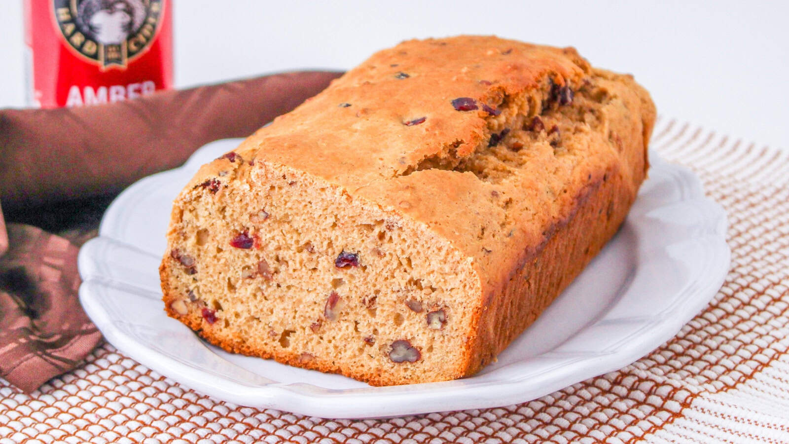 Loaf of bread with visible nuts and raisins on a white plate, placed on a textured surface next to a brown cloth and a can with partial label showing Amber.