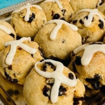 A tray of round scones with raisins, topped with icing crosses, on a silver platter.