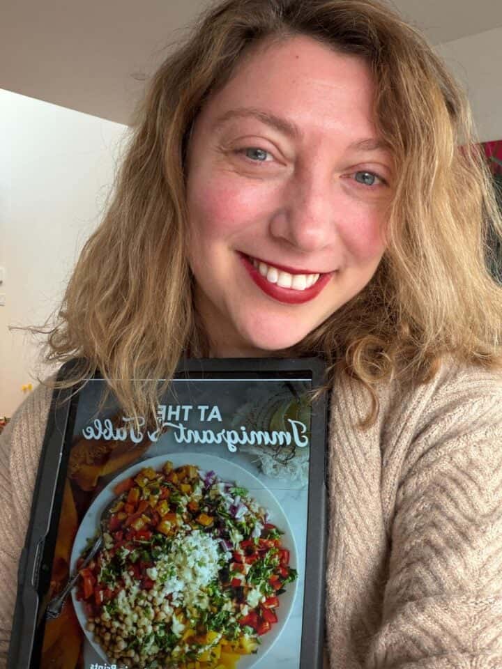 Smiling woman holds a tablet displaying a colorful salad and the words "At The Immigrant’s Table E-Book.
