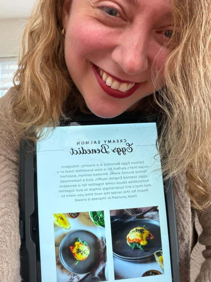 Smiling woman holding a tablet displaying a recipe for creamy salmon eggs benedict with food photos from At The Immigrant’s Table E-Book.