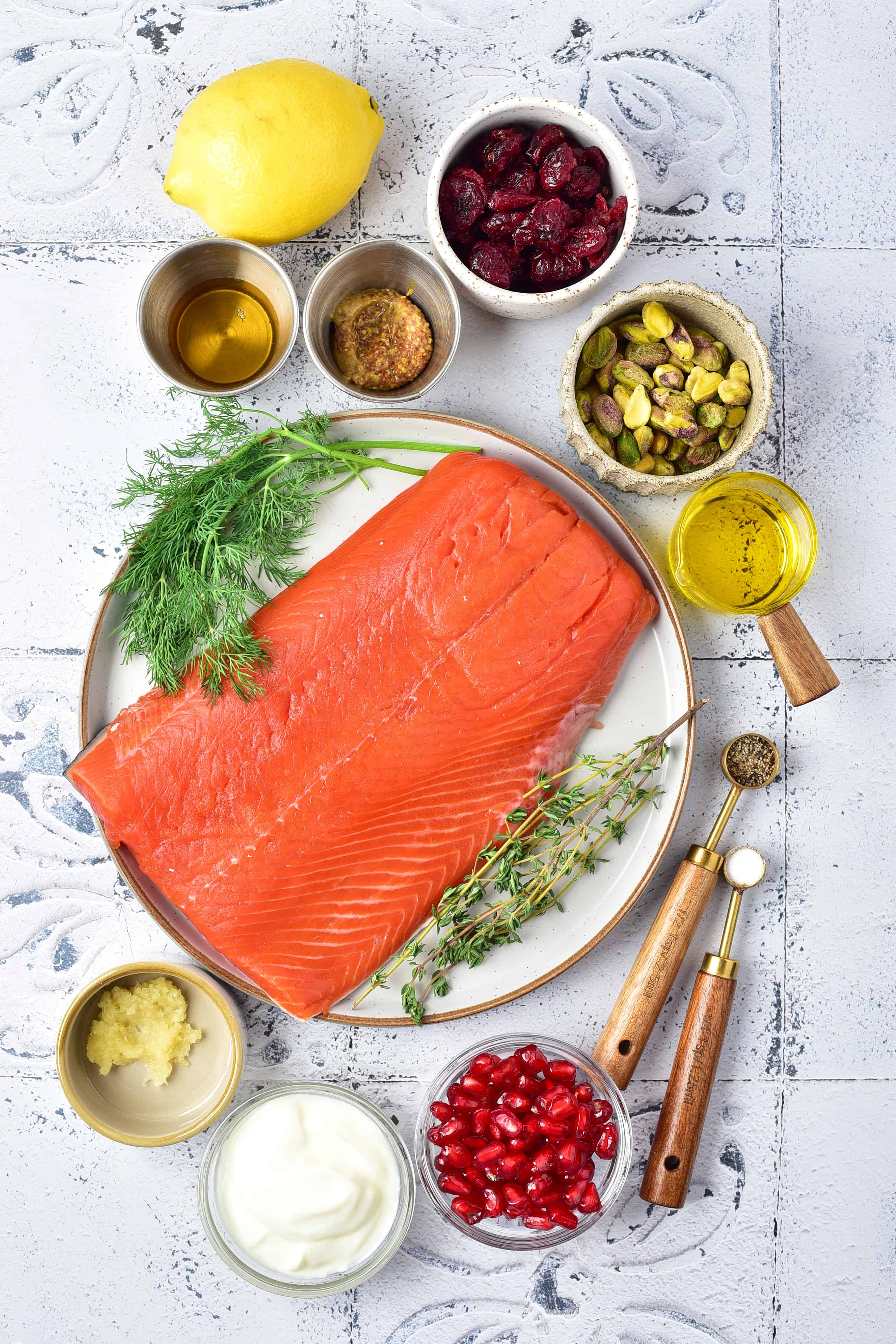 Fresh salmon fillet on a plate, surrounded by herbs, lemon, nuts, berries, mustard, oil, and sauce on a white table.