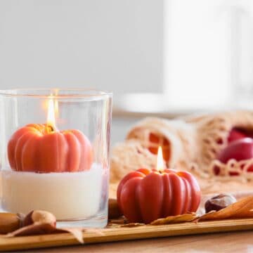 A lit pumpkin-shaped candle in a glass and another on a tray with autumn leaves and nuts—perfect for cozy pumpkin spice gifts.