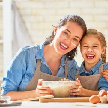 A smiling woman and girl in aprons mix dough together in a bright kitchen, flour on their faces, surrounded by kitchen tools for holiday baking.