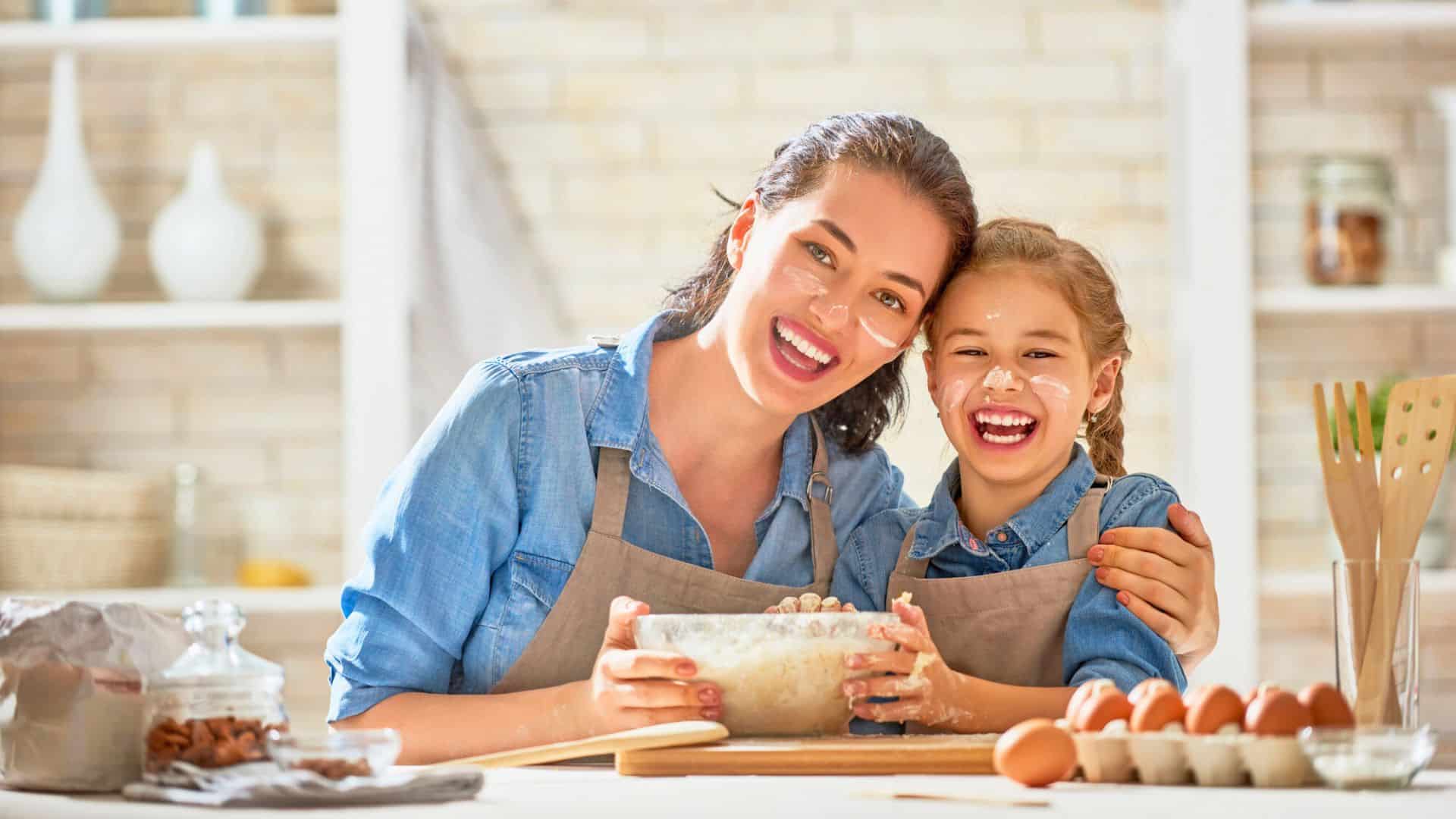 A smiling woman and girl in aprons mix dough together in a bright kitchen, flour on their faces, surrounded by kitchen tools for holiday baking.