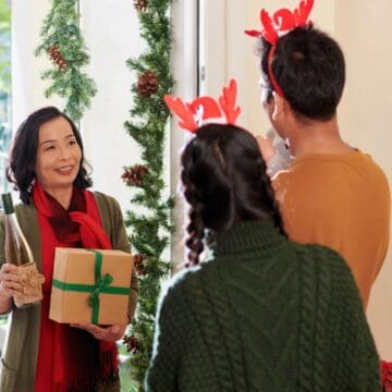 Woman holding a gift and wine greets two people with reindeer headbands at a festive holiday doorway, sharing thoughtful gifts for holiday hosts and entertainers.