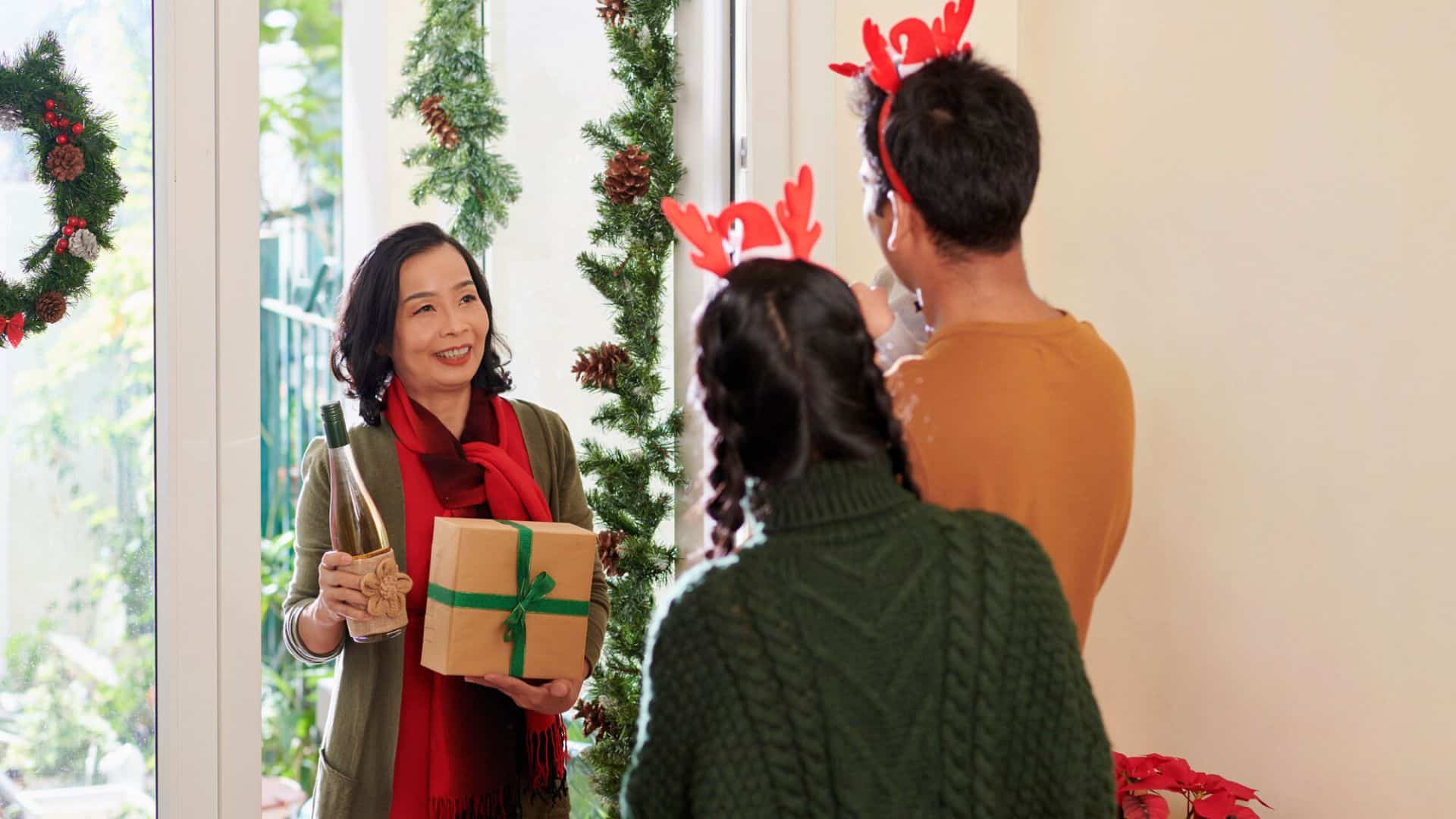 Woman holding a gift and wine greets two people with reindeer headbands at a festive holiday doorway, sharing thoughtful gifts for holiday hosts and entertainers.