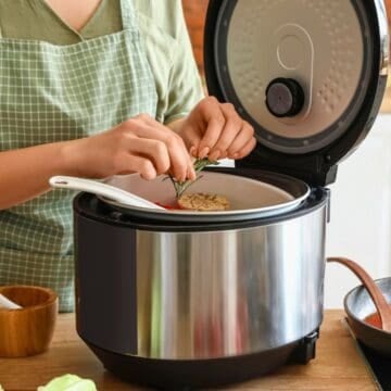 Person in green apron adding herbs to food in an electric cooker, using kitchen gadgets for holiday cooking, with fresh ingredients nearby.