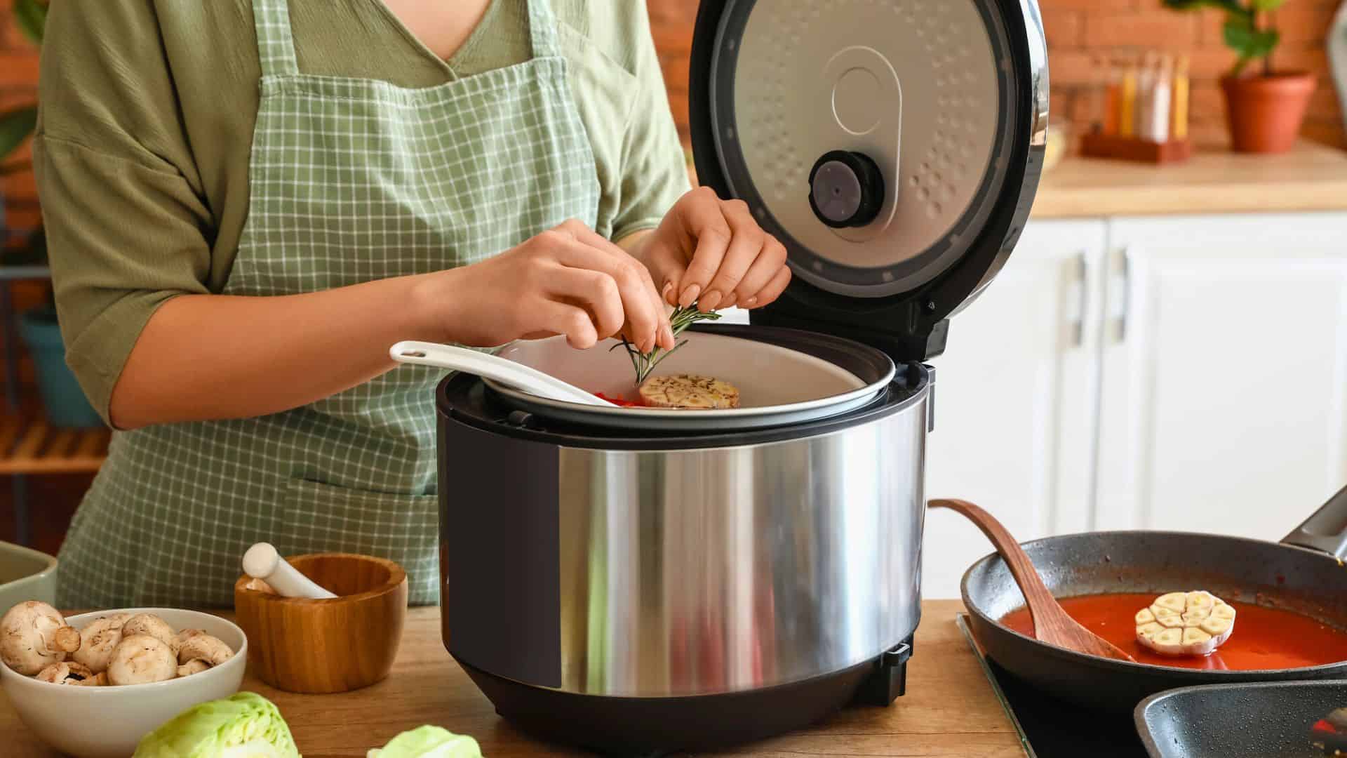 Person in green apron adding herbs to food in an electric cooker, using kitchen gadgets for holiday cooking, with fresh ingredients nearby.
