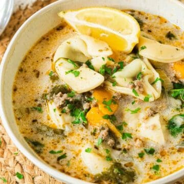 A bowl of creamy tortellini soup with spinach, lemon wedges, and herbs, served with a spoon.