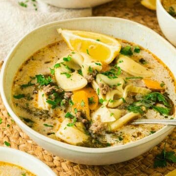 A bowl of creamy soup with vegetables, herbs, lemon wedges, and a spoon on a woven placemat.