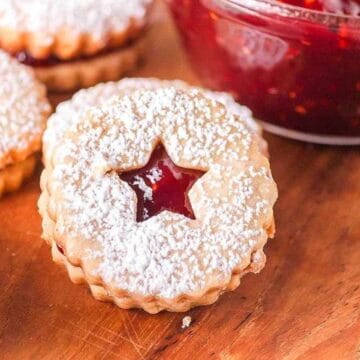 Round jam-filled cookies with powdered sugar and star-shaped cutouts on a wooden surface near a bowl of jam.
