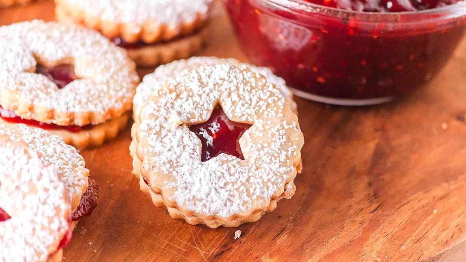 Round jam-filled cookies with powdered sugar and star-shaped cutouts on a wooden surface near a bowl of jam.