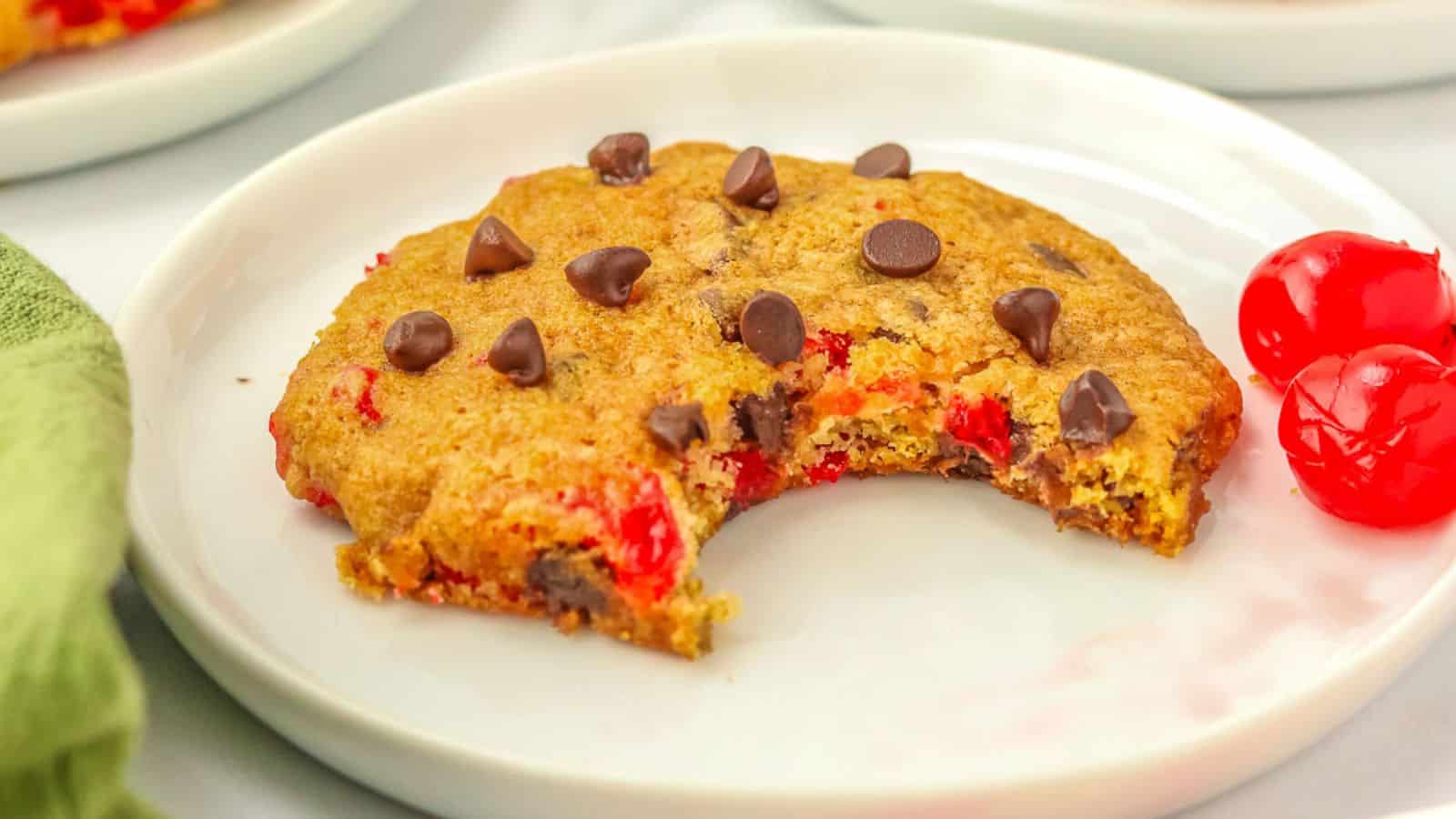 A cookie with a bite taken out, topped with chocolate chips, sits on a plate next to two cherries.