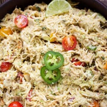 A bowl of creamy shredded chicken salad with cherry tomatoes, jalapeño slices, and lime on top.