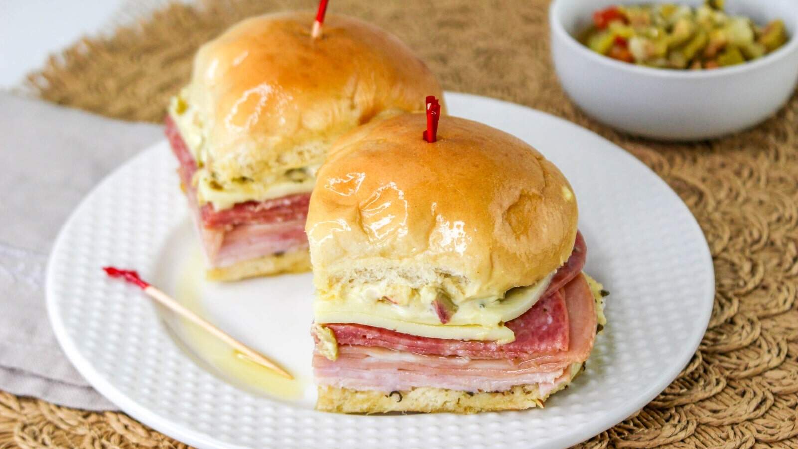 Two slider sandwiches with deli meats and cheese on a white plate, each secured with a toothpick. A small bowl of olives is in the background.