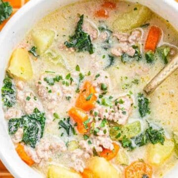 A bowl of creamy soup with ground meat, potatoes, carrots, kale, and herbs on an orange tiled surface.