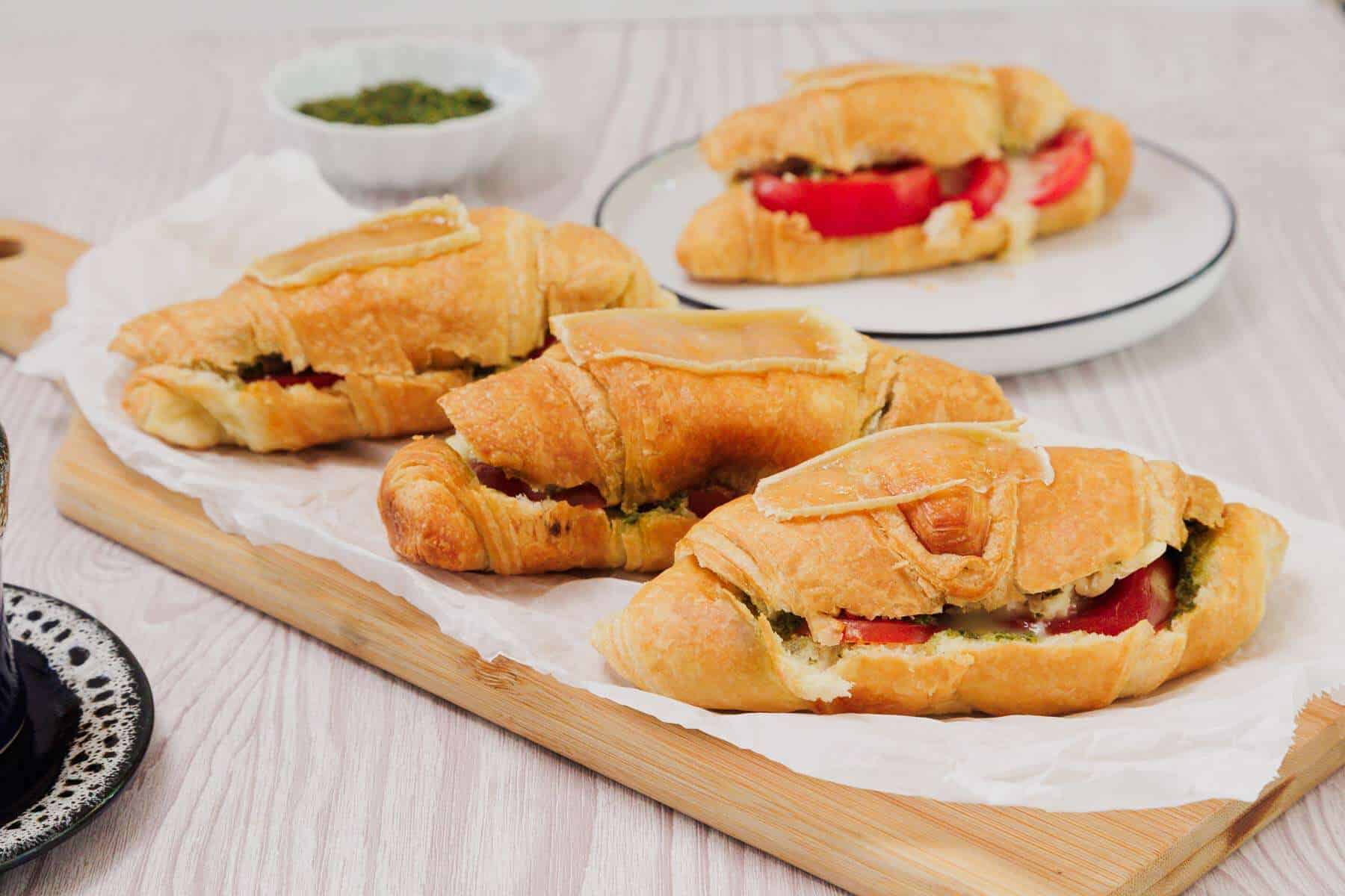 Three croissant sandwiches with tomato and cheese on a wooden board, with more sandwiches on a plate in the background.