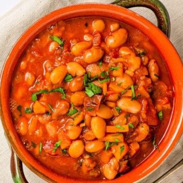 A bowl of bean stew in tomato sauce, garnished with herbs, on a beige cloth next to a spoon.