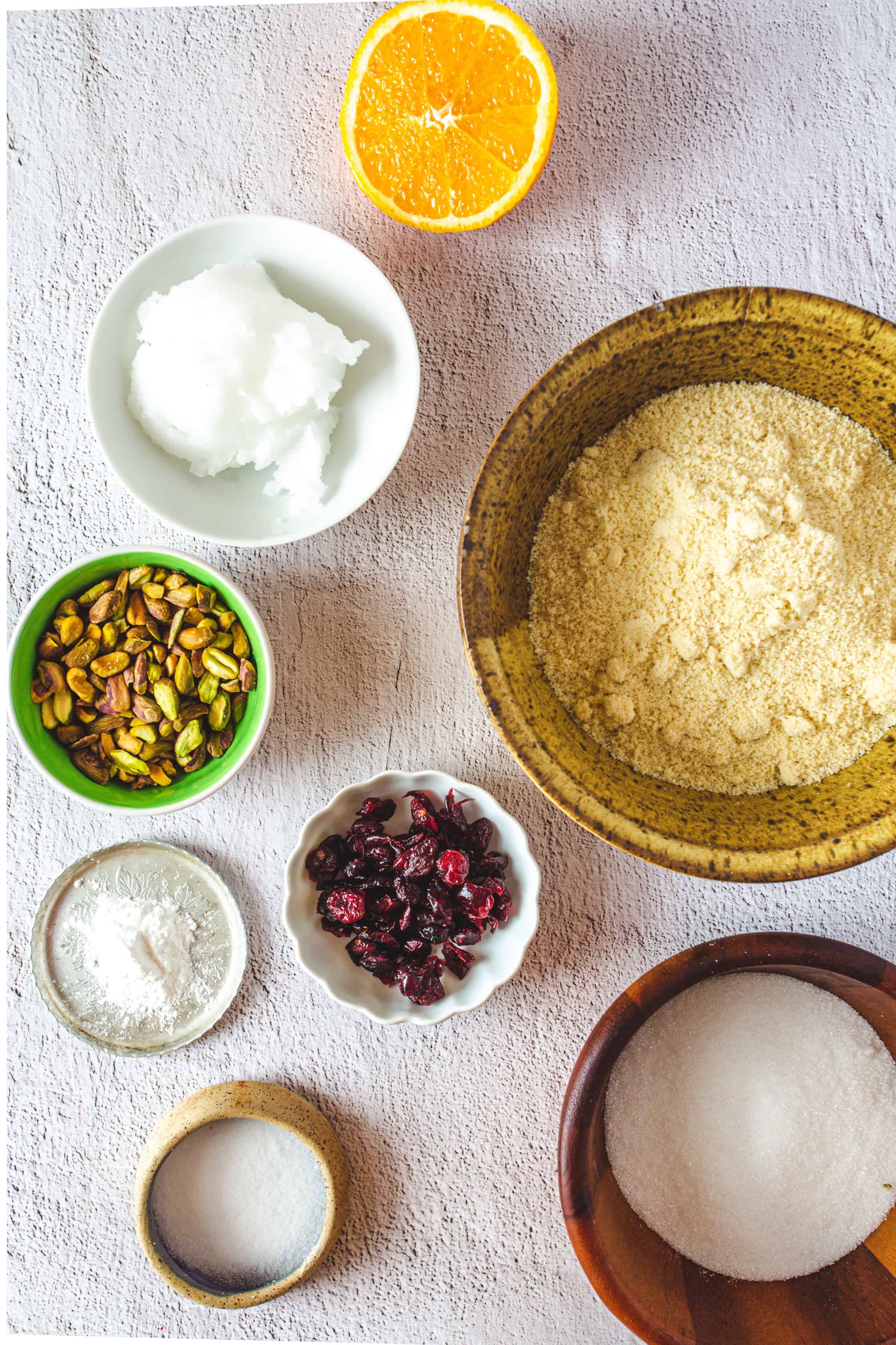 Top view of bowls with ingredients for Pistachio Gluten-Free Biscotti, including almond flour, sugar, coconut oil, pistachios, dried cranberries, baking powder, salt, and an orange half.