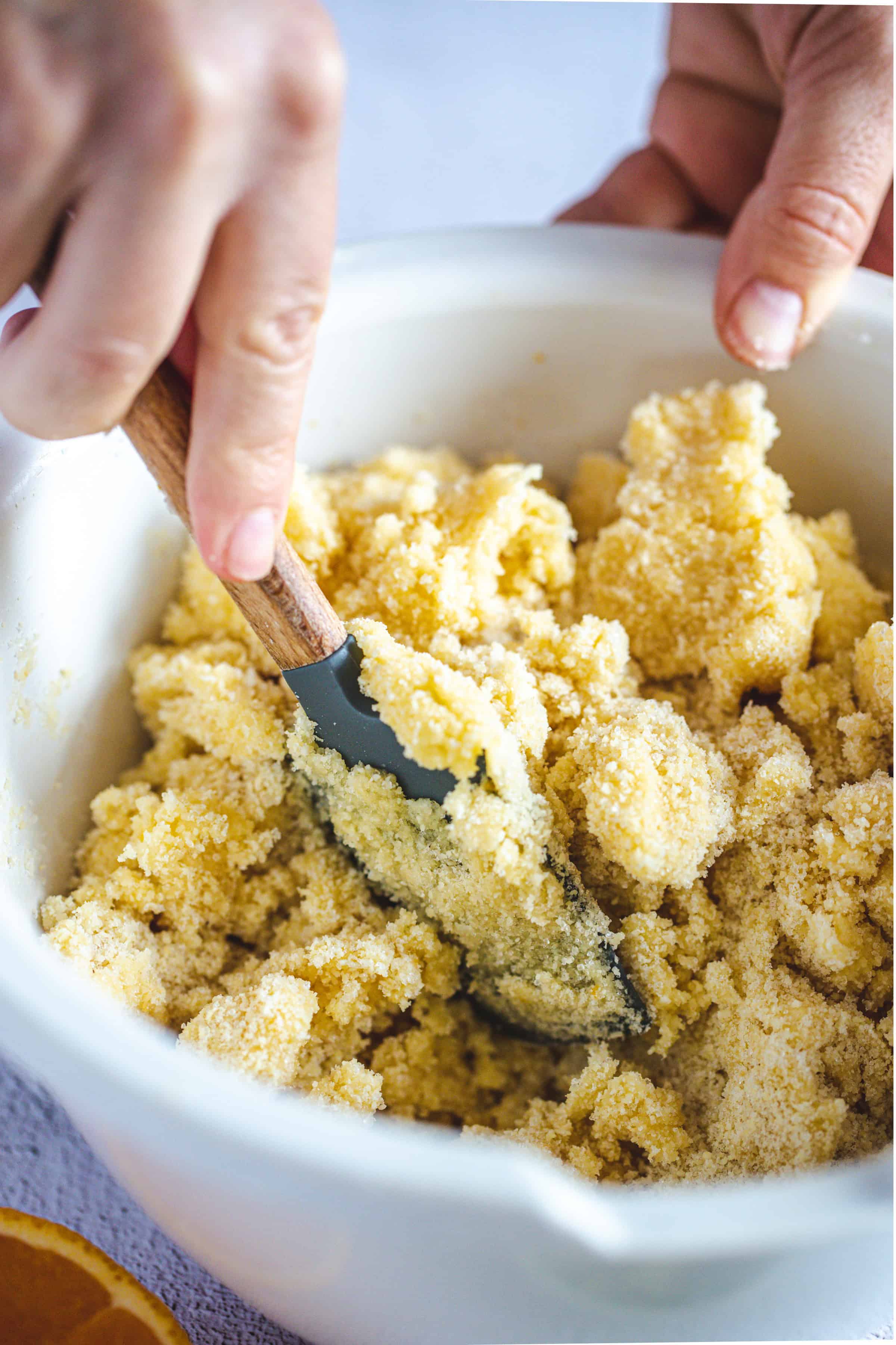 A hand mixes coarse, yellowish sugar in a white bowl with a spatula, preparing the base for Pistachio Gluten-Free Biscotti.
