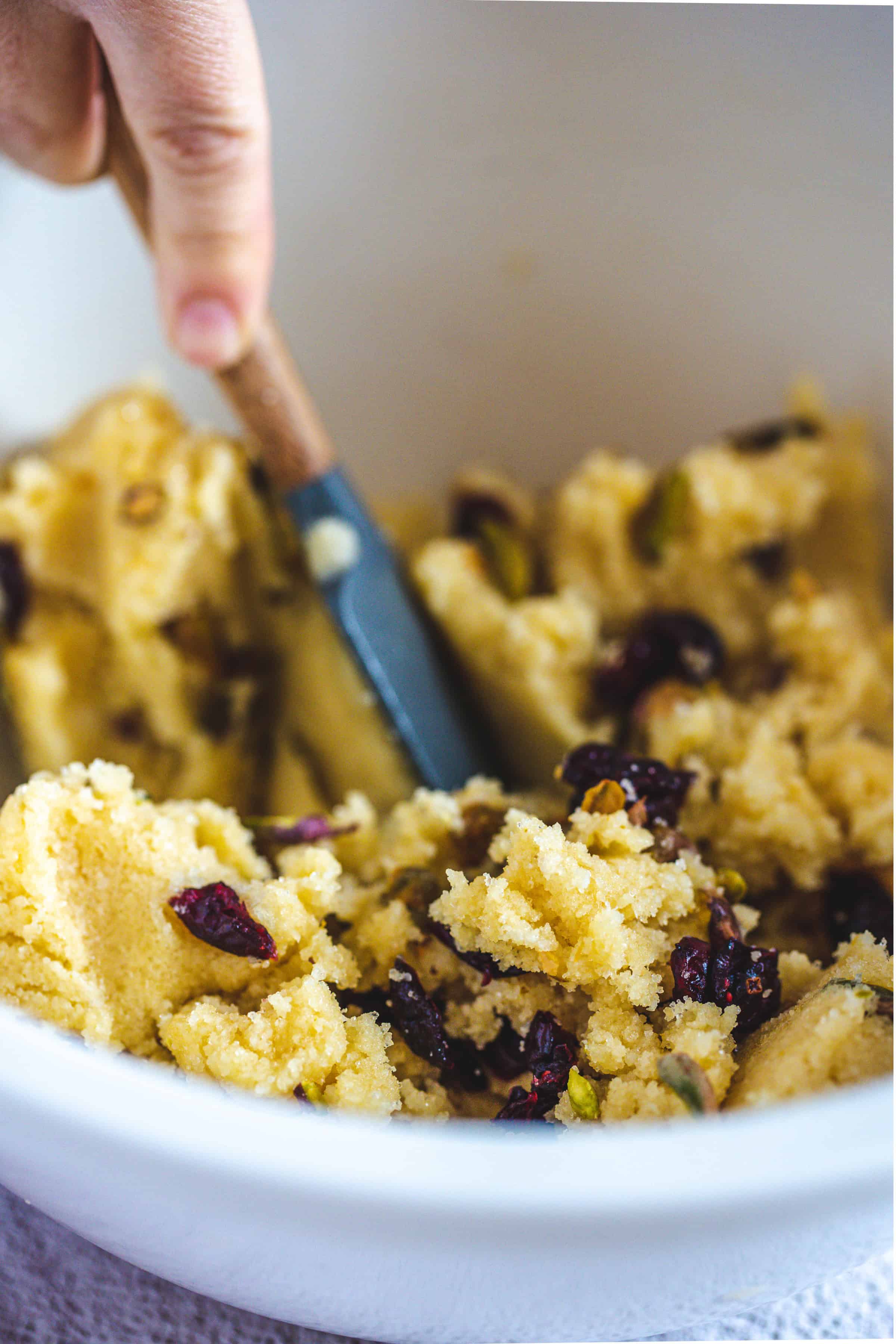 Hand mixing dough for Pistachio Gluten-Free Biscotti with dried cranberries and nuts in a white bowl using a spatula.
