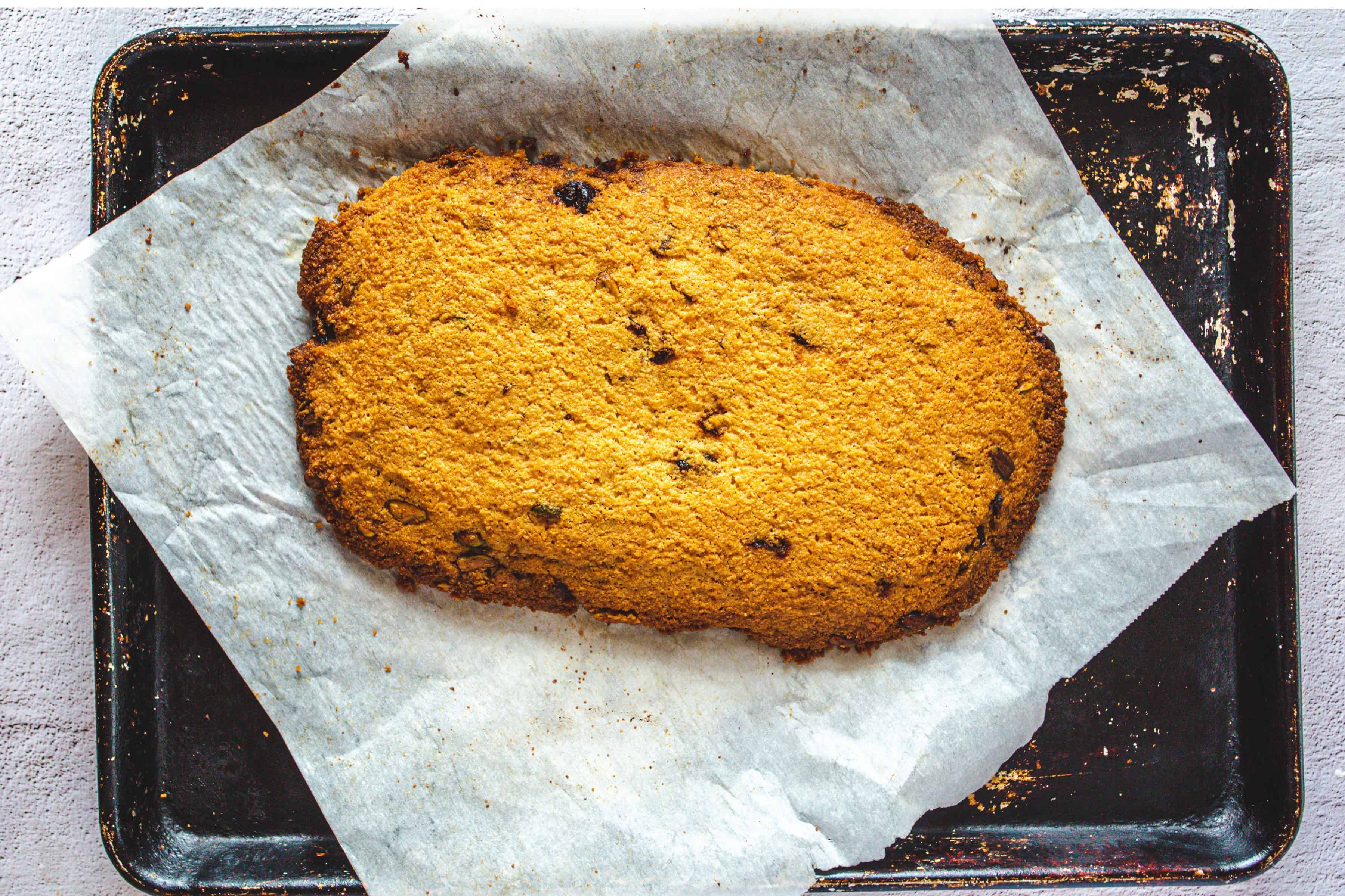 A large, rustic golden-brown Pistachio Gluten-Free Biscotti rests on parchment paper atop a worn baking tray.