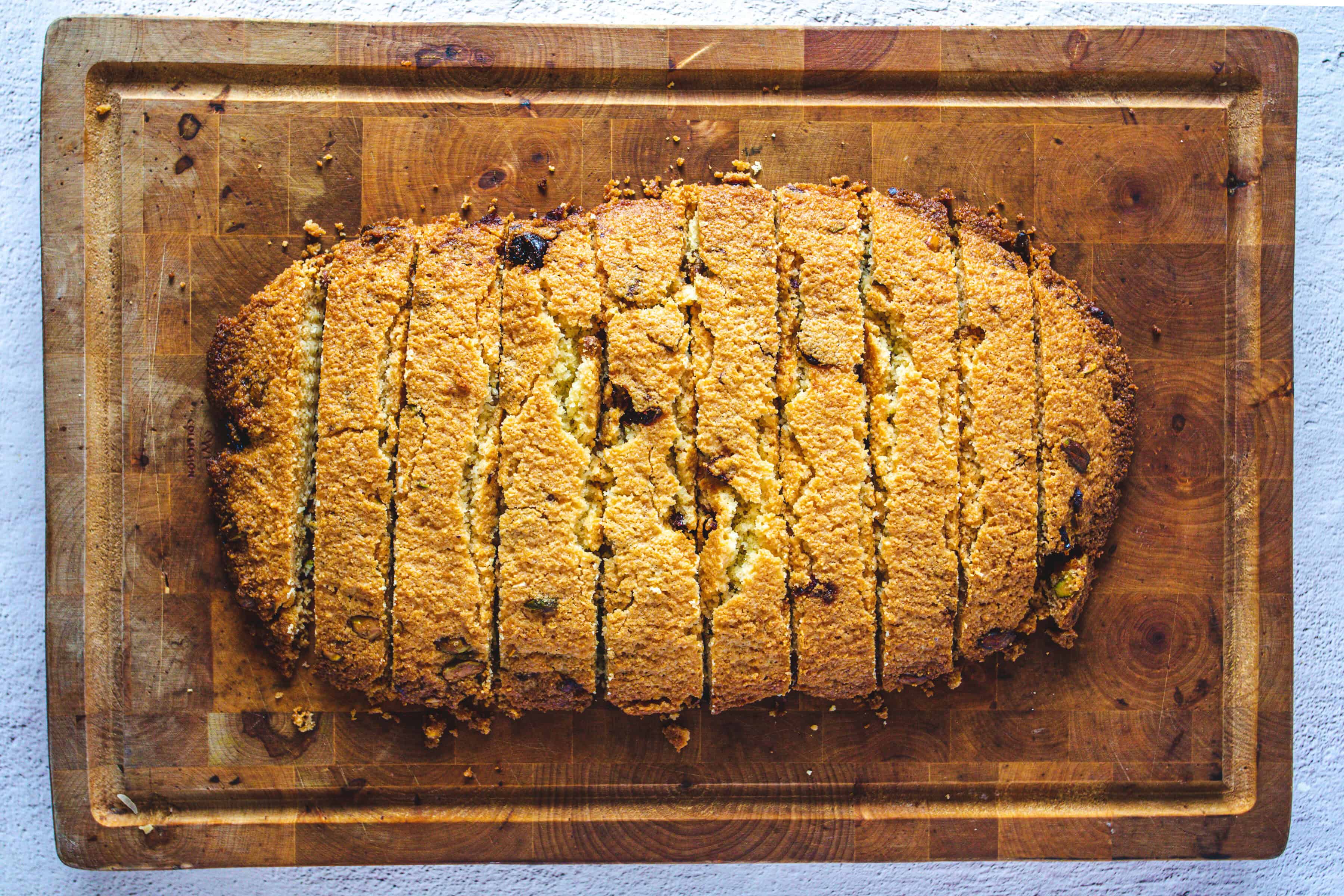 A large, golden-brown loaf of bread sliced on a wooden cutting board, next to crunchy Pistachio Gluten-Free Biscotti.