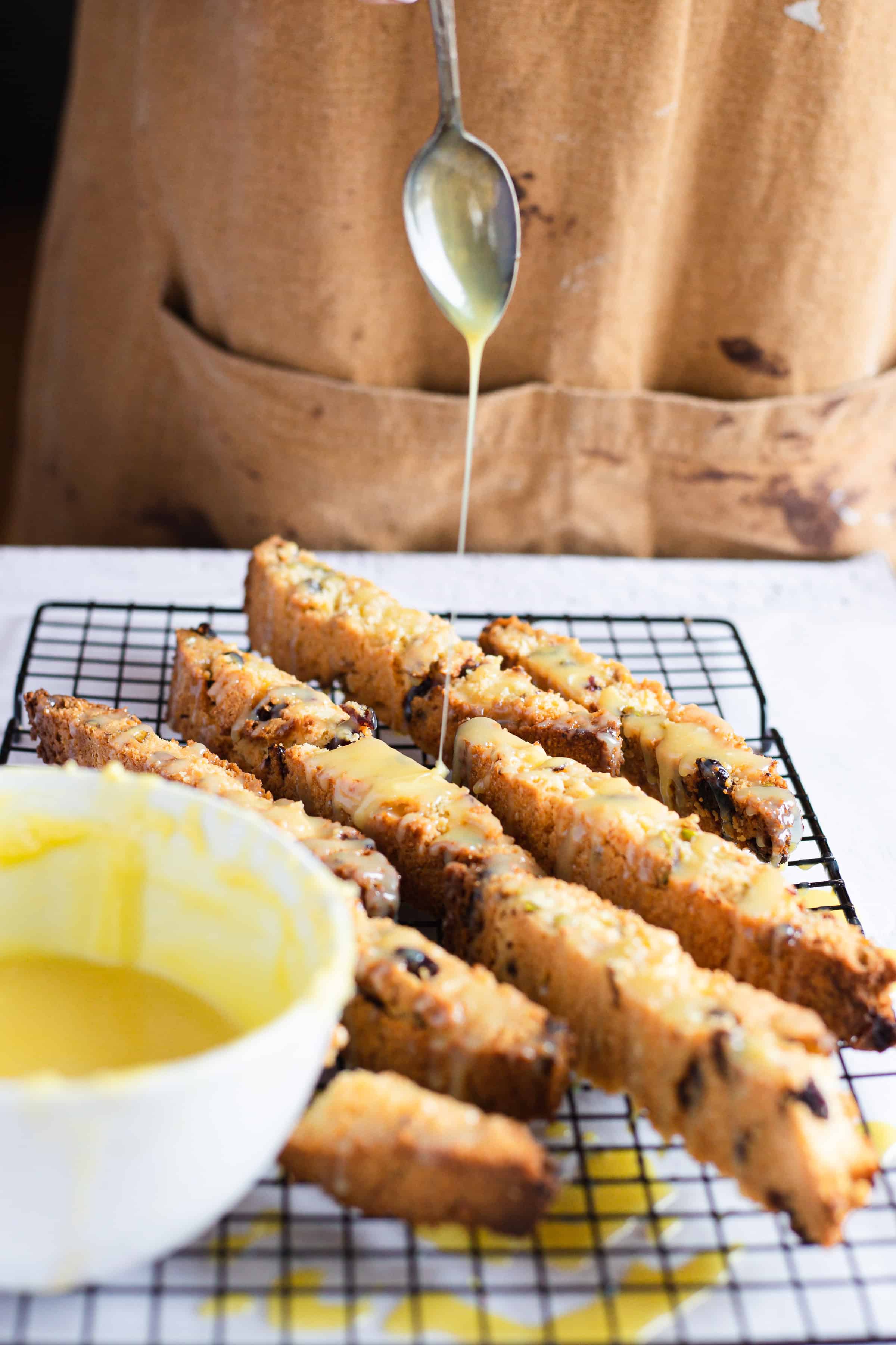 A person drizzles icing over Pistachio Gluten-Free Biscotti on a cooling rack, with a bowl of icing nearby.