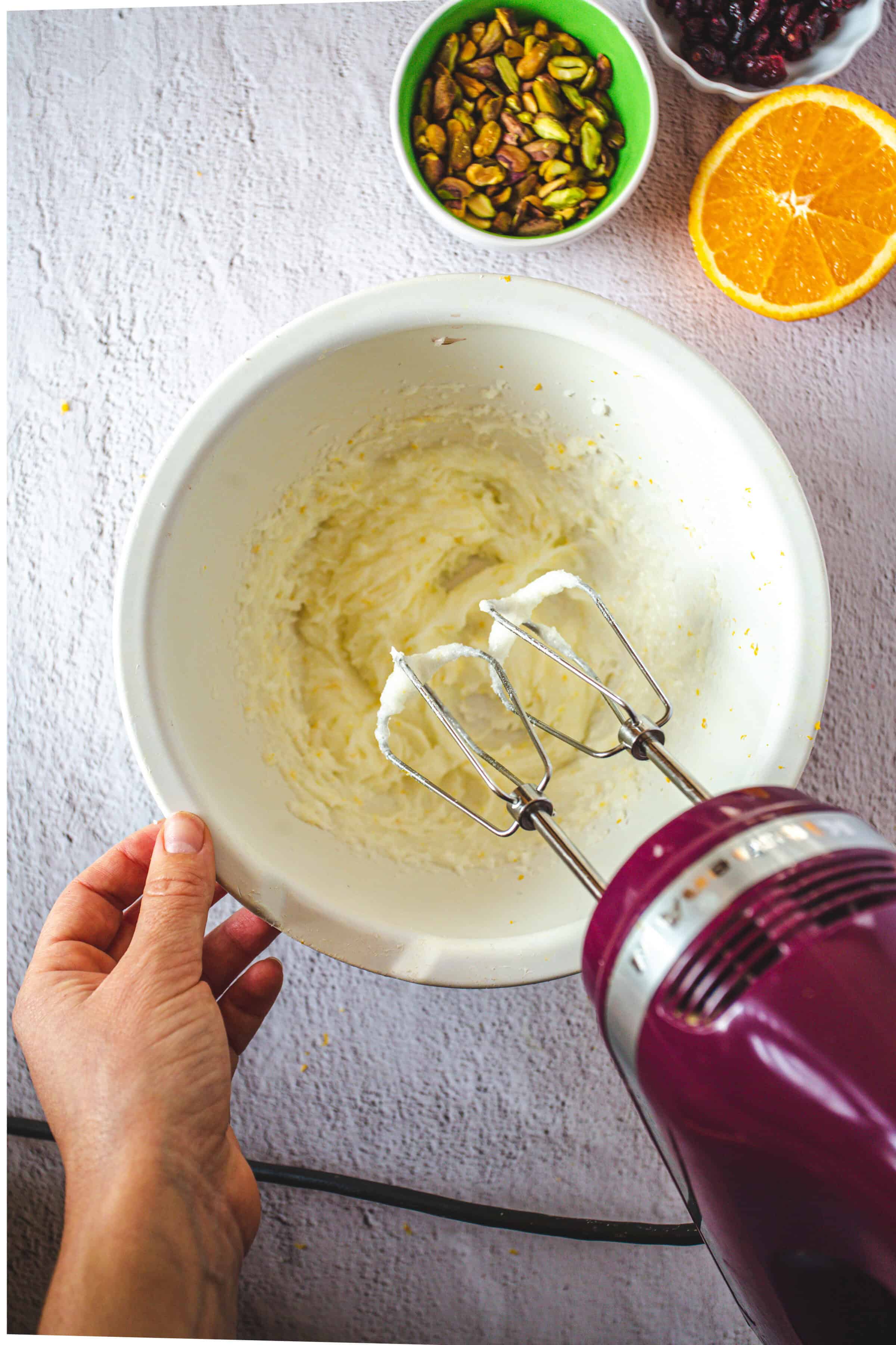 A hand holds a bowl with whipped mixture for Pistachio Gluten-Free Biscotti, an electric mixer, pistachios, orange, and berries nearby.