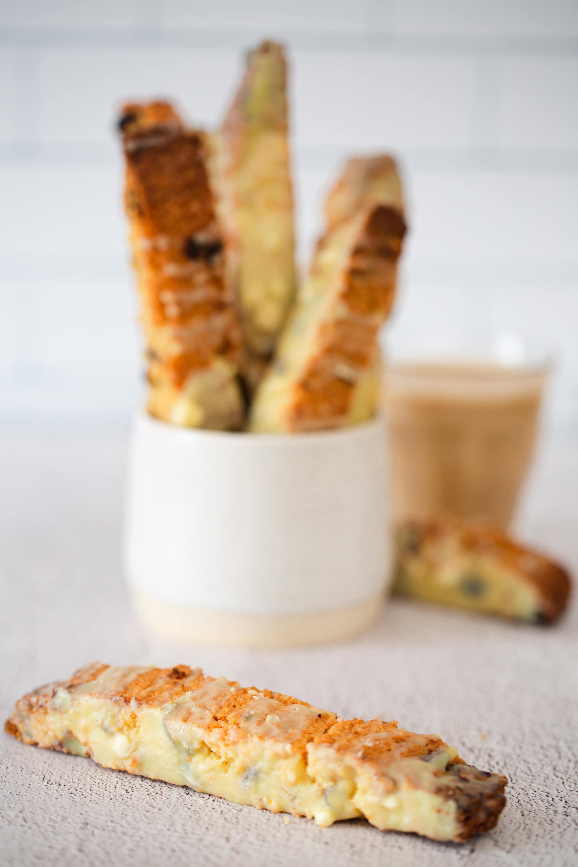 A white cup holds several Pistachio Gluten-Free Biscotti; one biscotti lies on the table in front, with a coffee cup in the background.