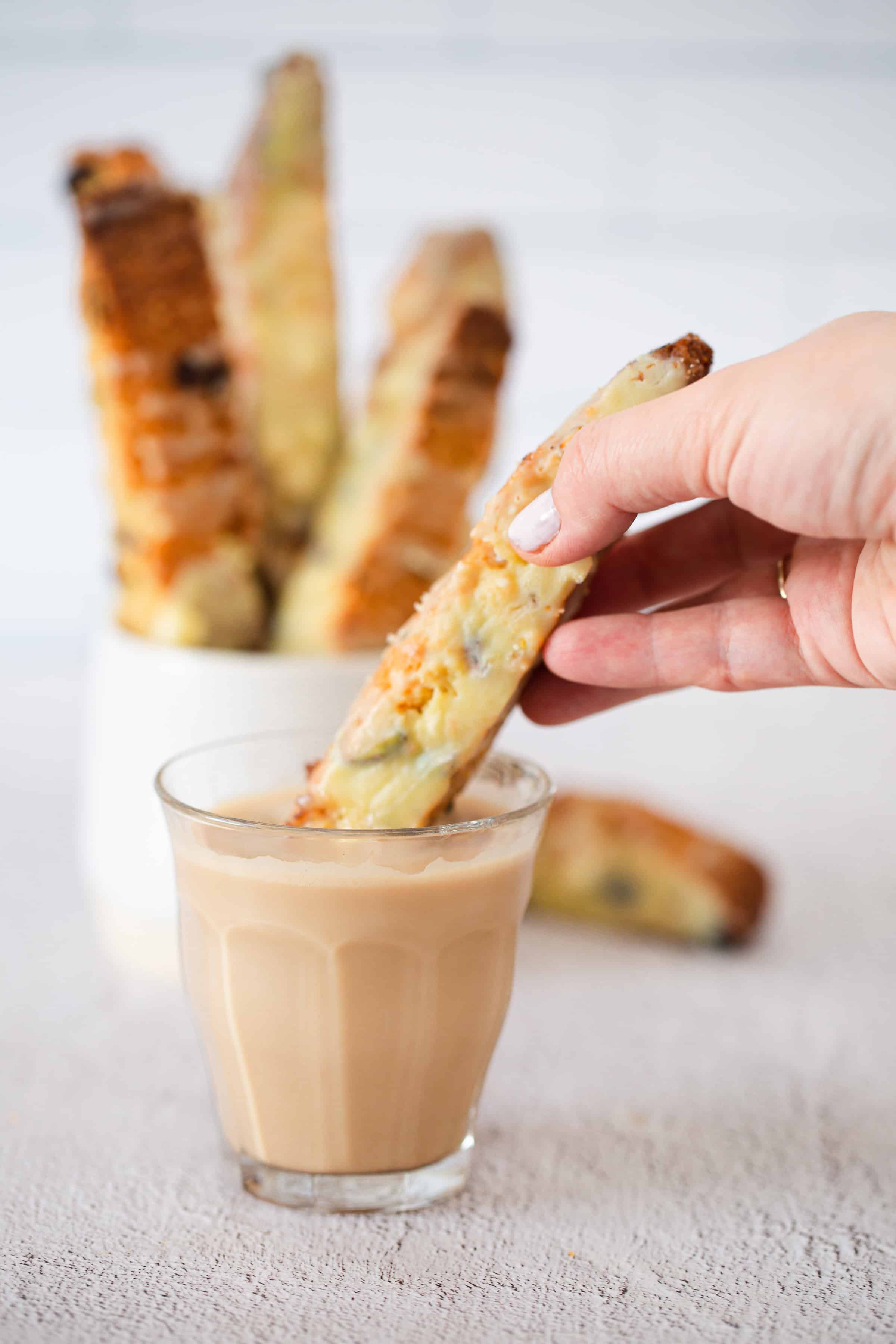 A hand dips a Pistachio Gluten-Free Biscotti into a glass of coffee, with more biscotti in a jar in the background.