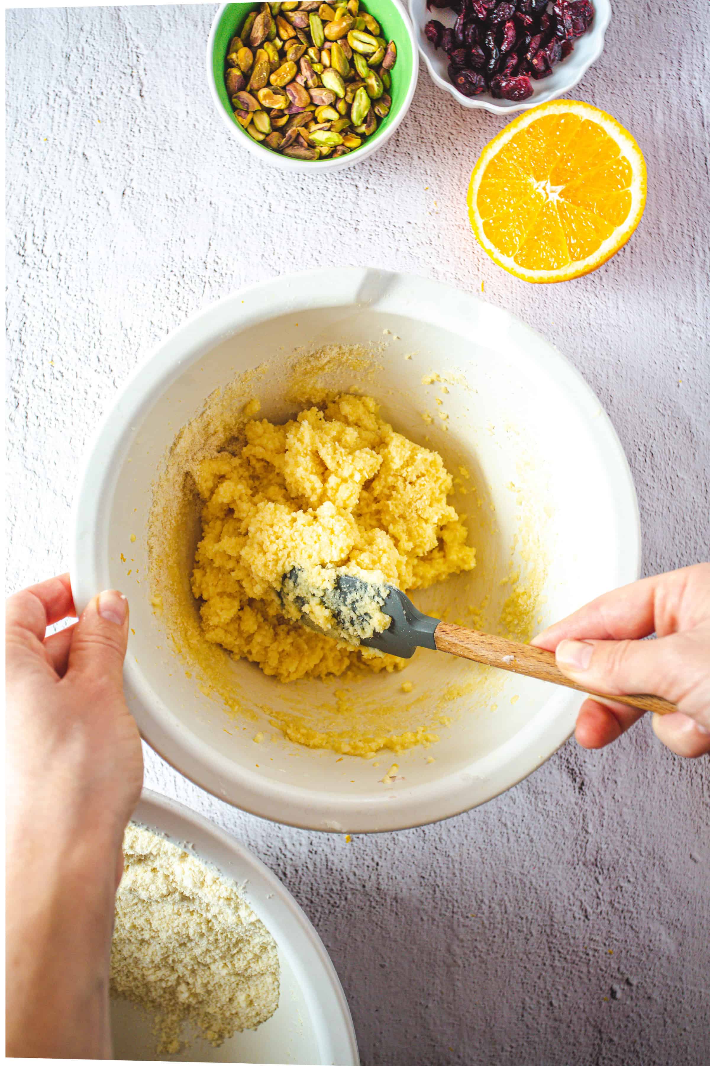 Hands mixing a yellow dough in a bowl, surrounded by pistachios, cranberries, orange, and almond meal-perfect ingredients for delicious Pistachio Gluten-Free Biscotti.