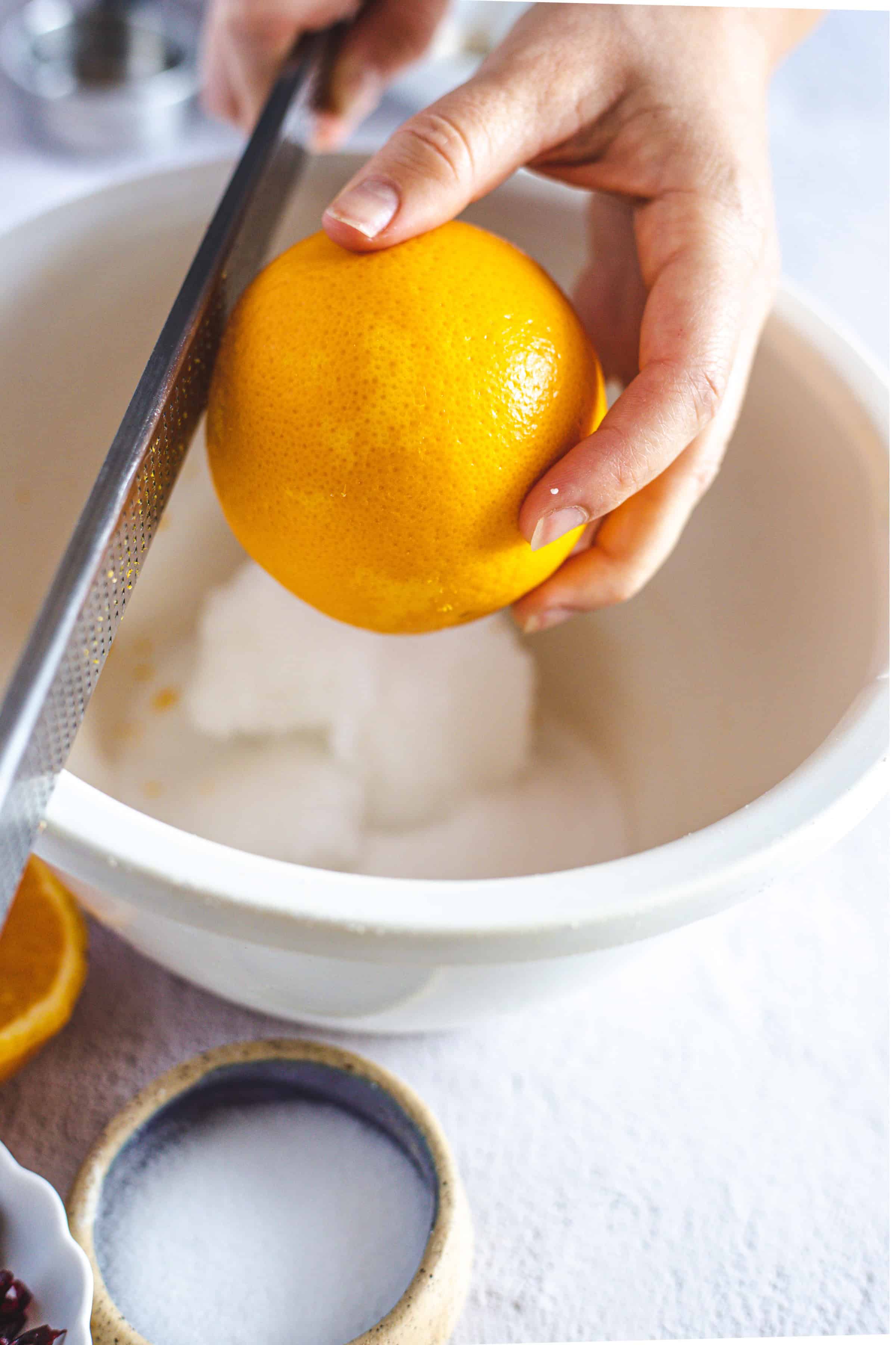 Hand zesting an orange over a white bowl with sugar inside, creating a fresh burst of citrus flavor for Pistachio Gluten-Free Biscotti, on a light background.