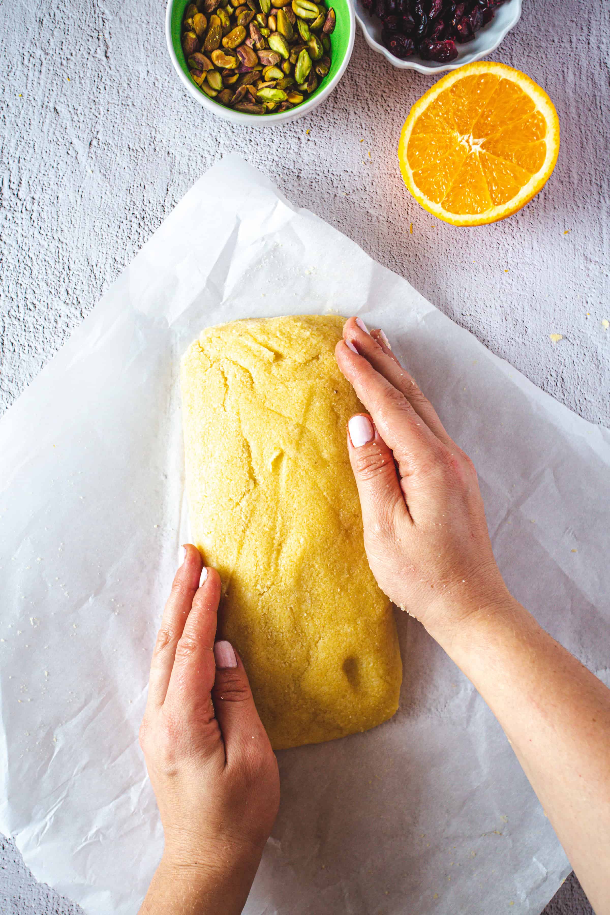 Hands shaping yellow dough on parchment paper, with bowls of pistachios, cranberries, and a halved orange nearby-perfect for making Pistachio Gluten-Free Biscotti.
