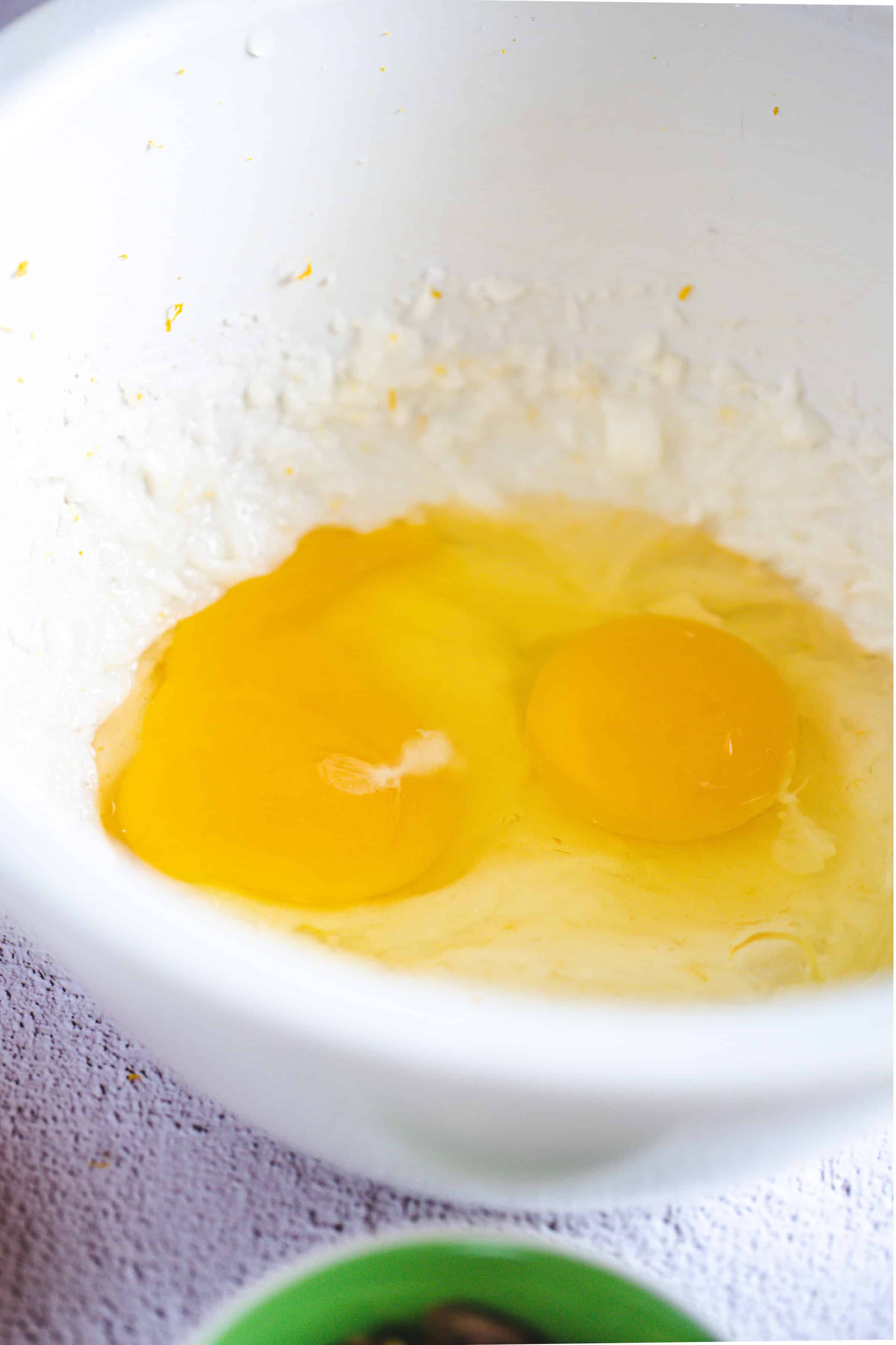 Two raw eggs cracked into a white mixing bowl with some Pistachio Gluten-Free Biscotti batter around the edges.