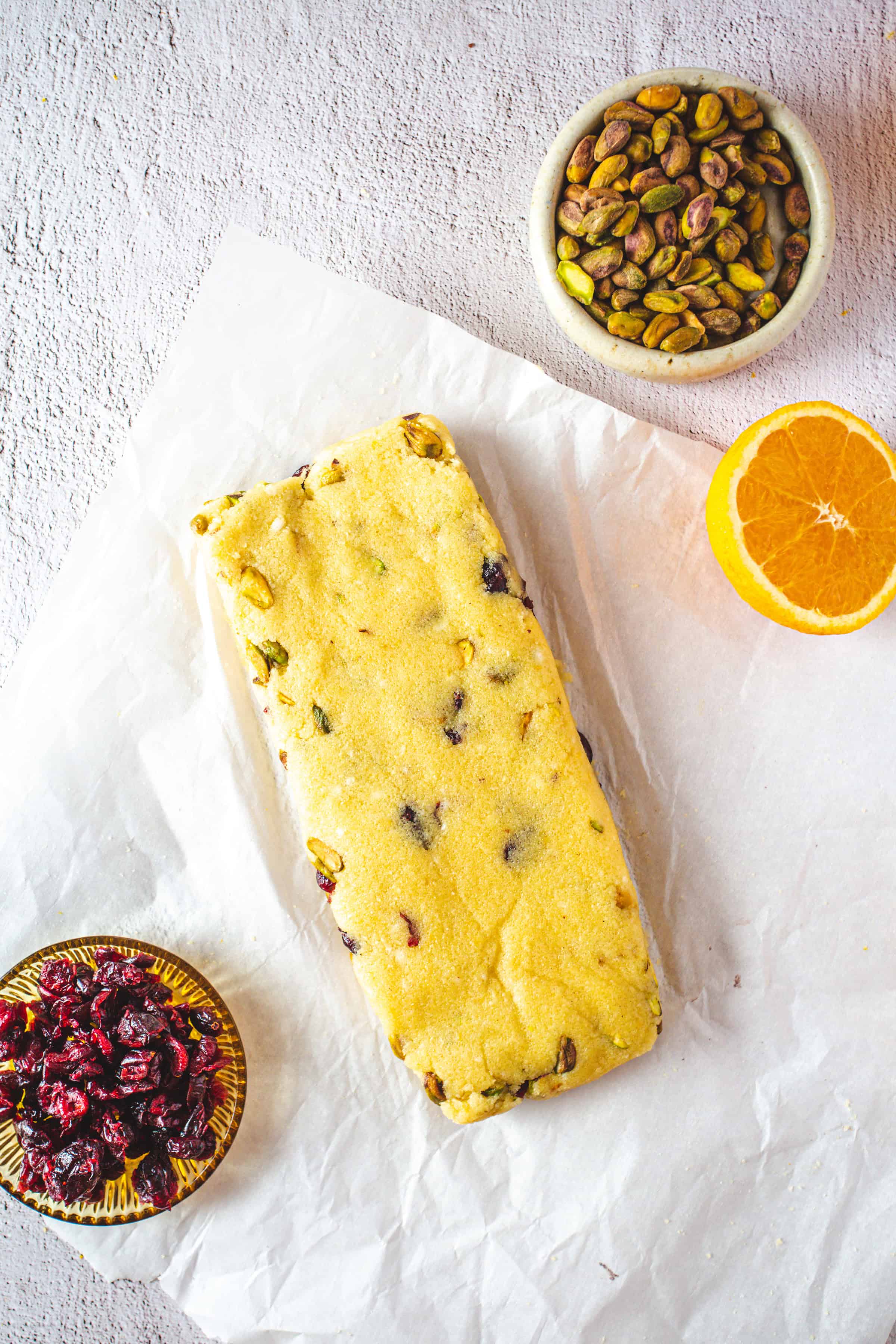 A loaf of Pistachio Gluten-Free Biscotti dough with cranberries sits on parchment, next to a bowl of pistachios, cranberries, and an orange half.