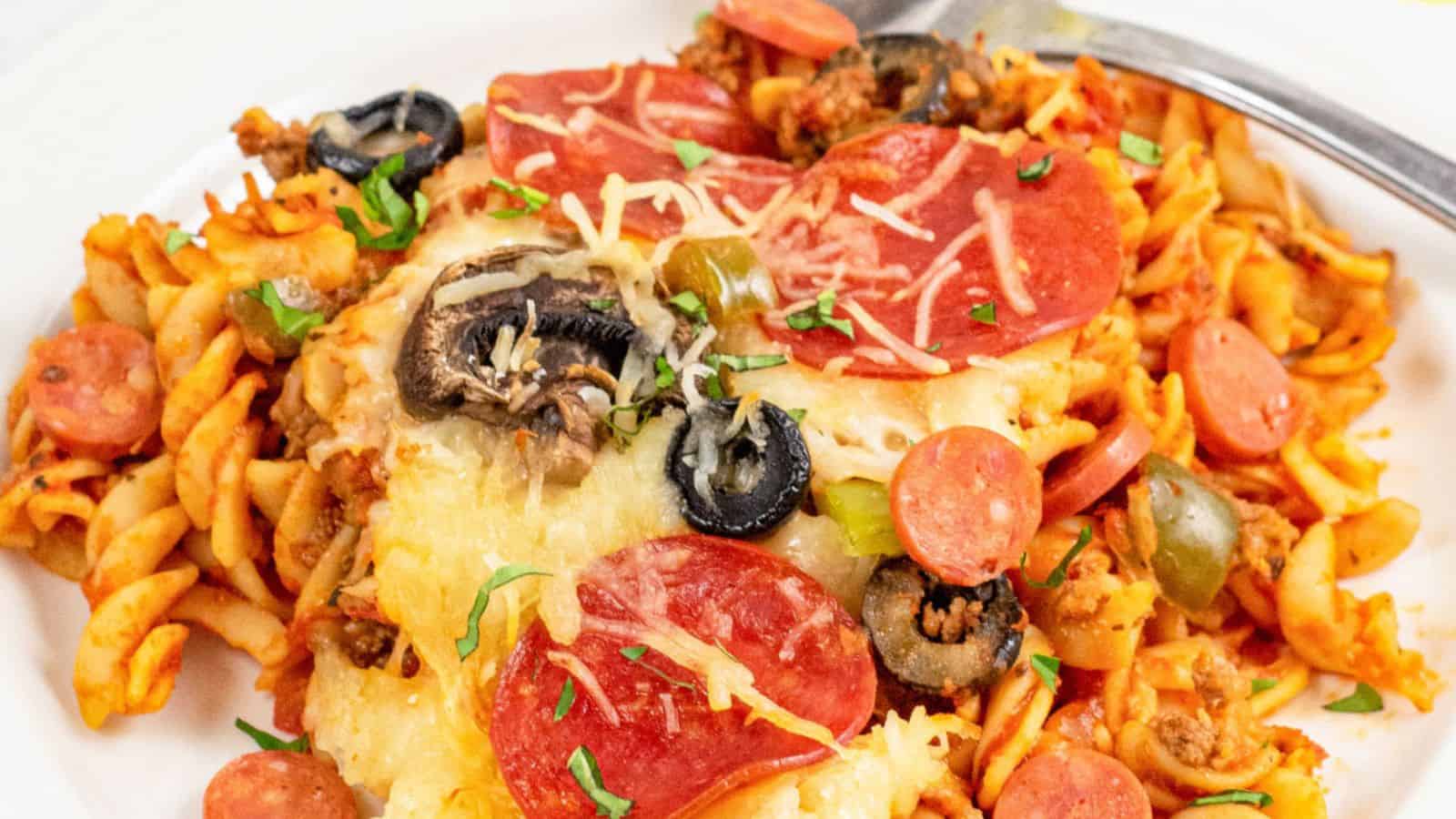 A plate of rotini pasta covered in tomato sauce, topped with pepperoni slices, olives, mushrooms, and melted cheese. Garnished with chopped herbs, this quick dish is served with a fork on the side.