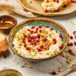 Bowl of creamy dip topped with pomegranate seeds and chopped pistachios, with bread slices nearby.