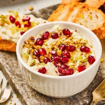 A bowl of whipped feta dip topped with pomegranate seeds and pistachios, served with toasted bread slices.