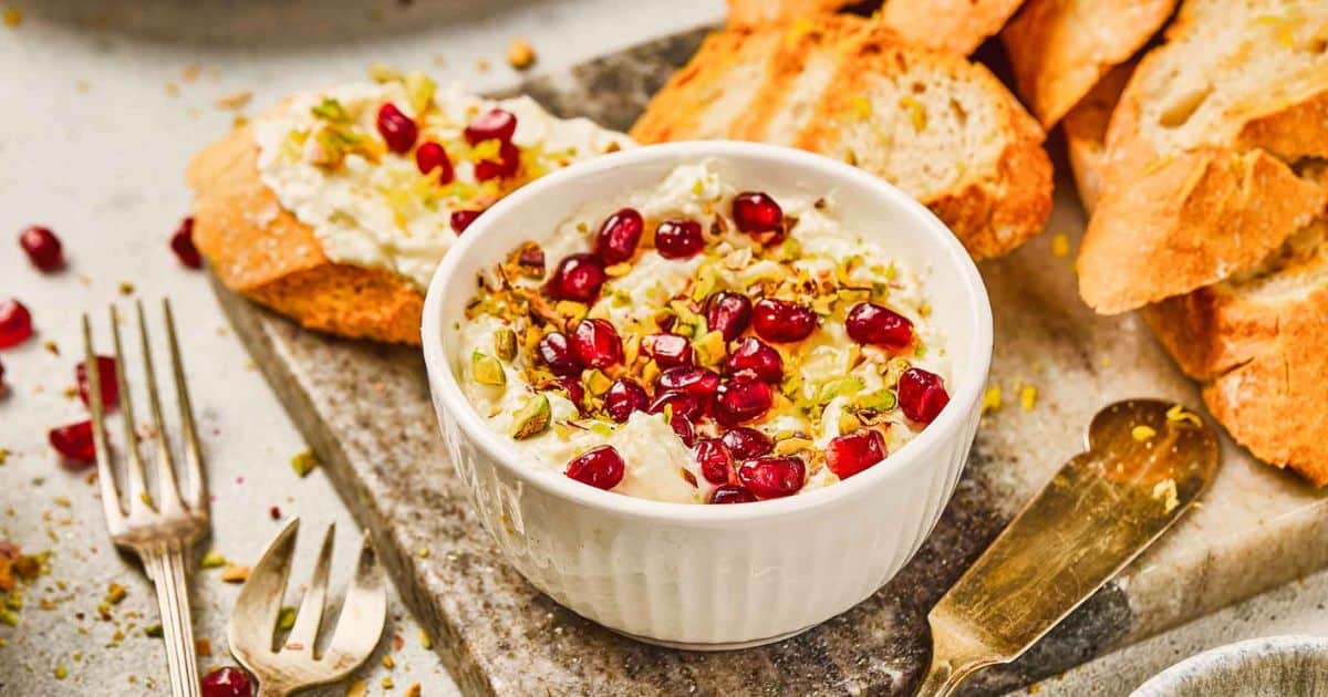 A bowl of whipped feta dip topped with pomegranate seeds and pistachios, served with toasted bread slices.