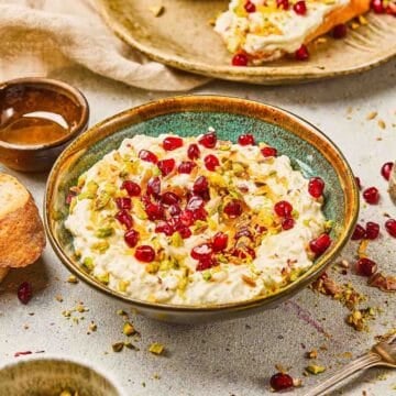 Bowl of creamy dip topped with pomegranate seeds and chopped pistachios, with bread slices nearby.