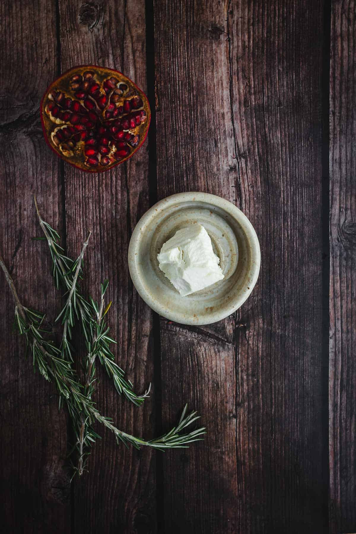 Bowl of pomegranate seeds, goat cheese, and rosemary sprigs on a rustic wooden surface.