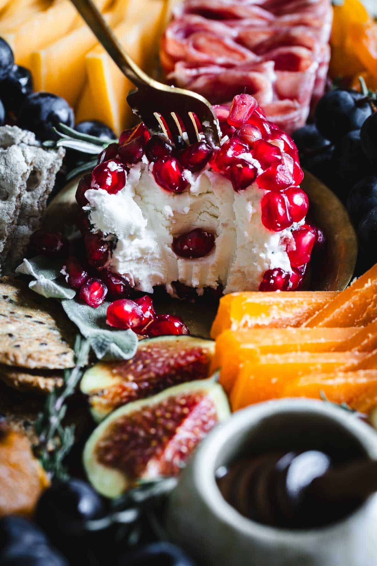 A fork dips into a cheese ball covered in pomegranate seeds, surrounded by figs, crackers, and sliced cheese.