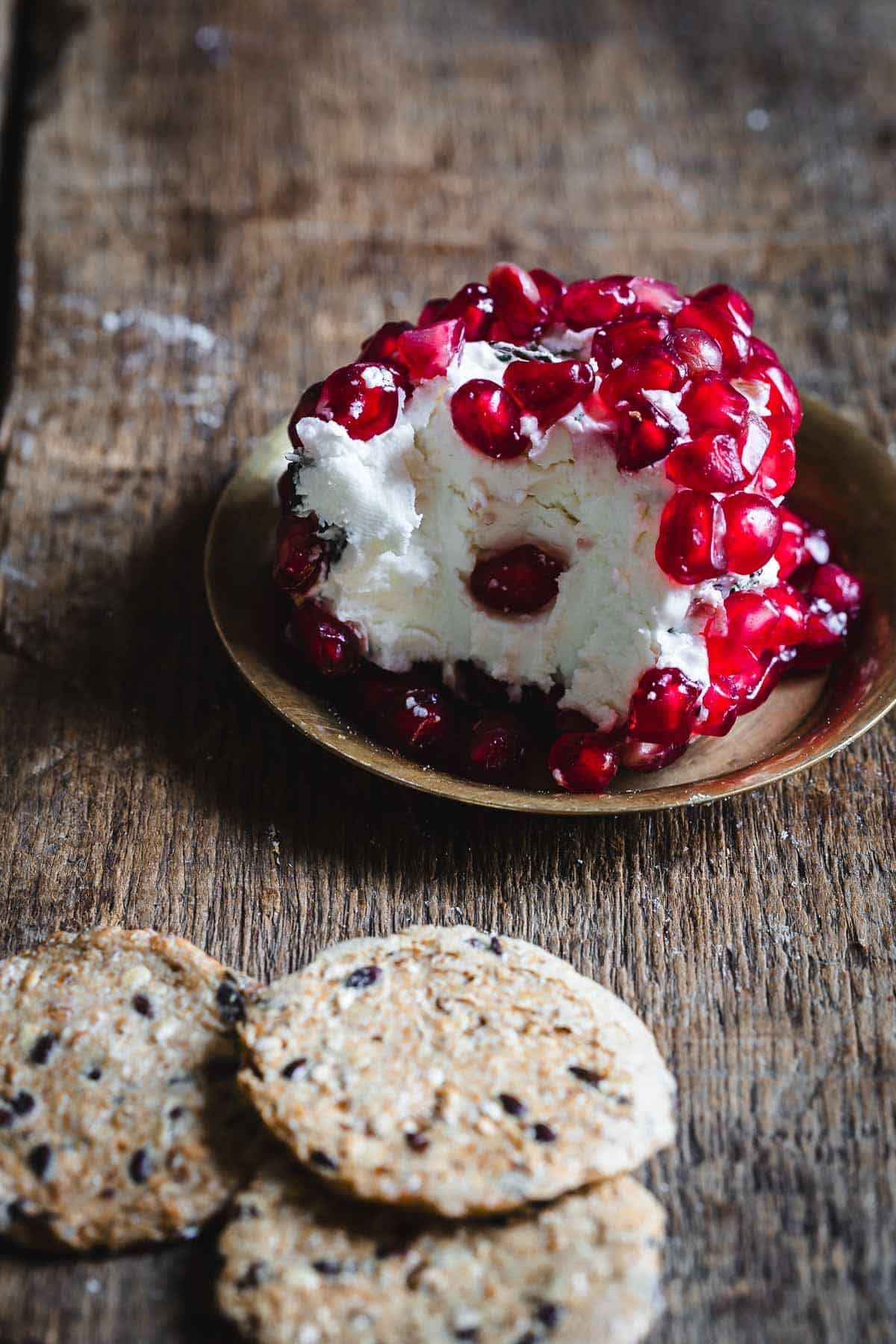 Goat cheese topped with pomegranate seeds on a brass plate, with crackers on a rustic wooden surface.