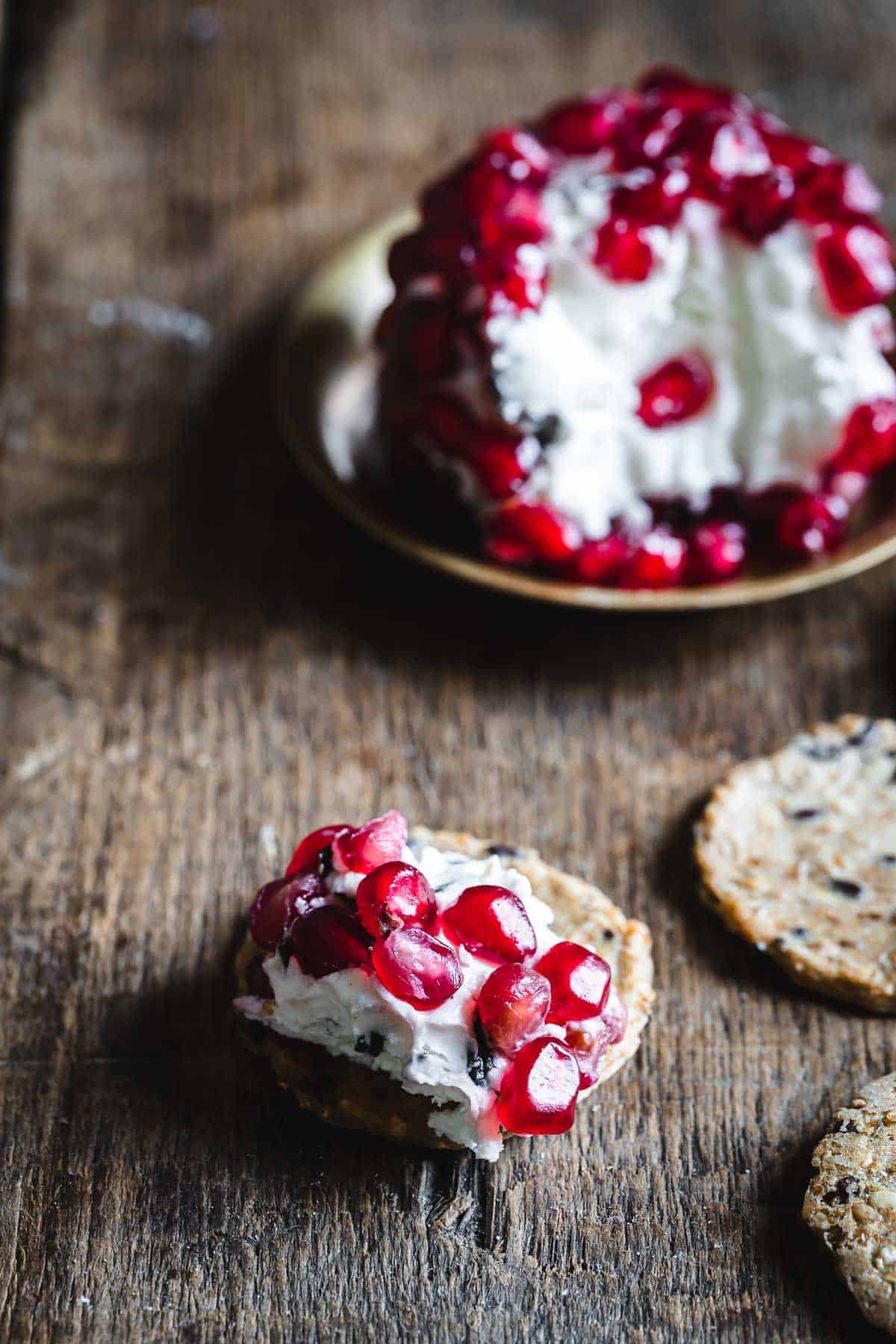 A seeded cracker topped with creamy cheese and pomegranate seeds on a rustic wooden surface.