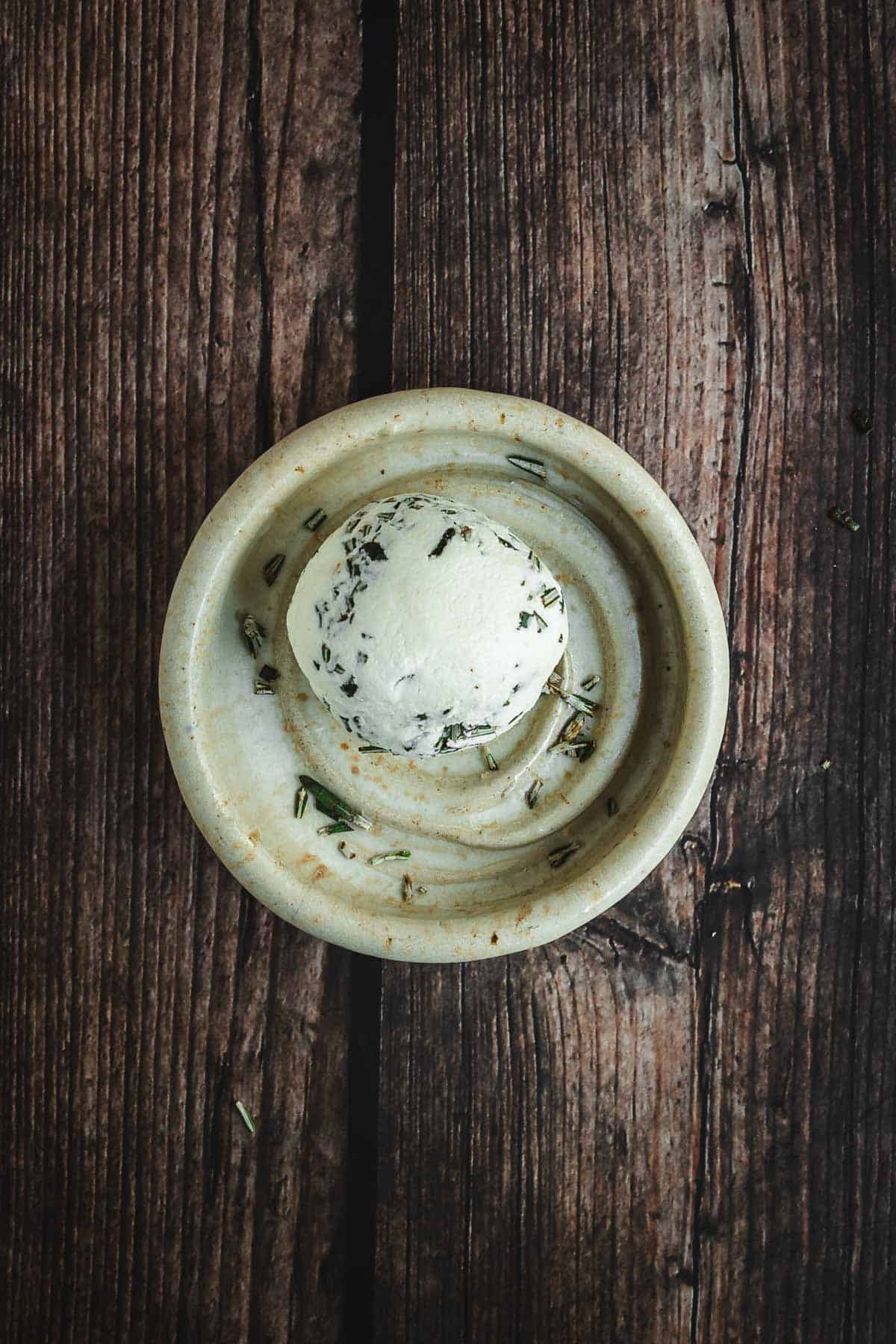 A festive Christmas Cheese Ball with fresh herbs sits on a rustic ceramic plate, set atop a weathered wooden surface.