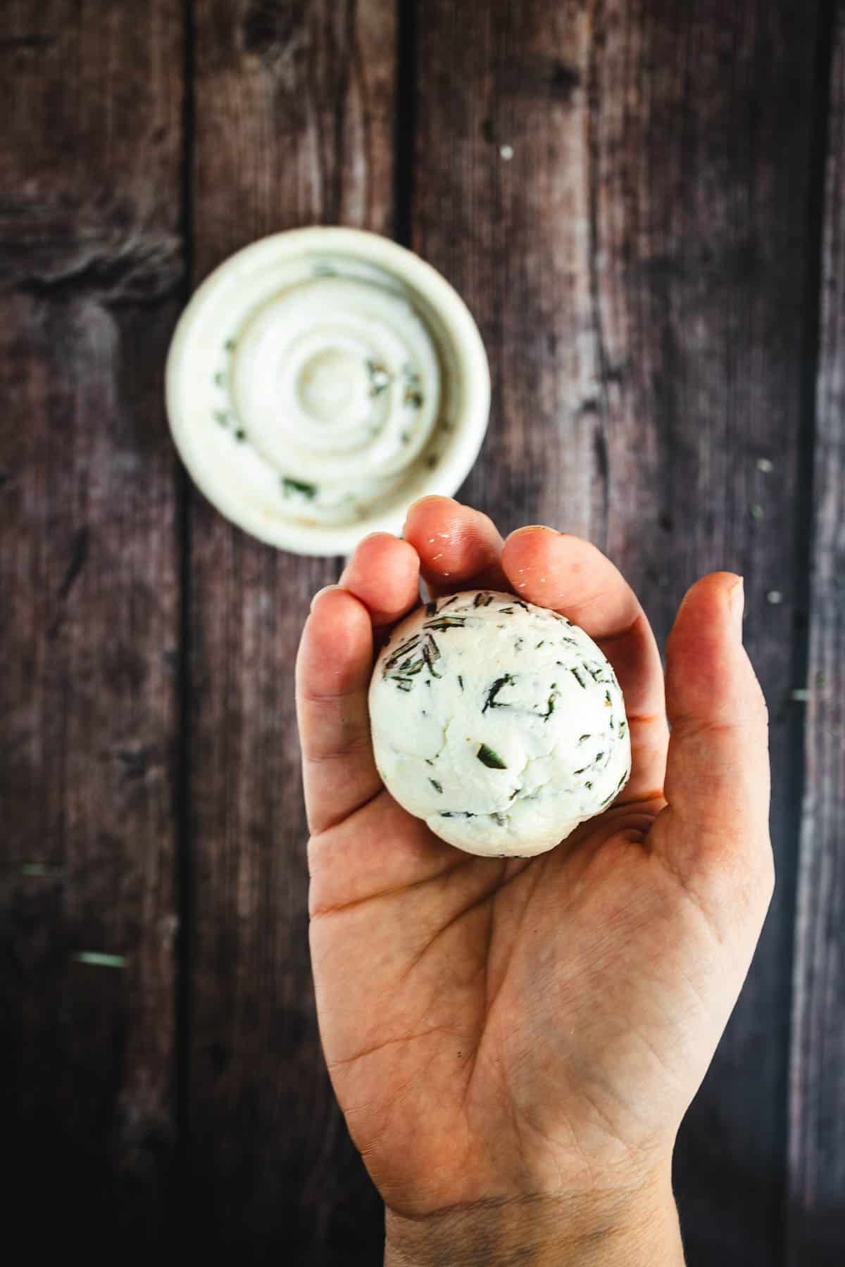 A hand holds a ball of herbed cheese above a small white dish on a wooden table.