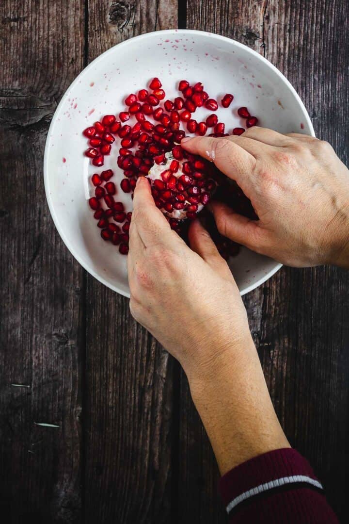 Hands removing pomegranate seeds over a white bowl on a dark wooden table.