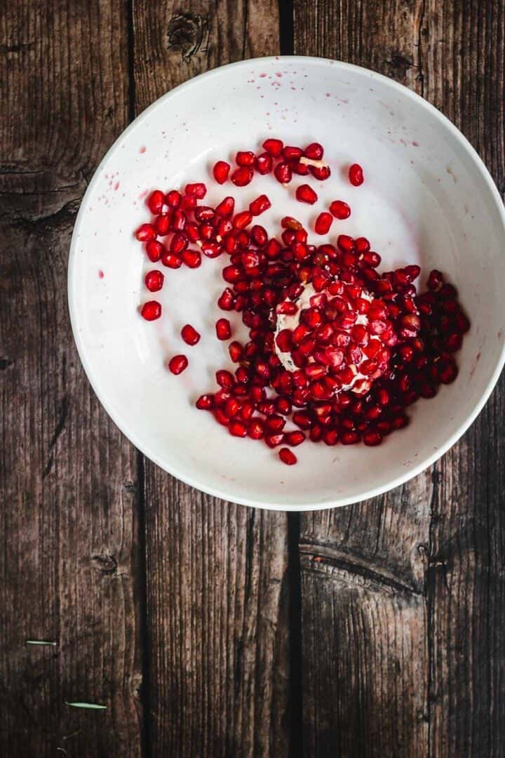 A white bowl with pomegranate seeds on a rustic wooden surface, some seeds scattered inside the bowl.