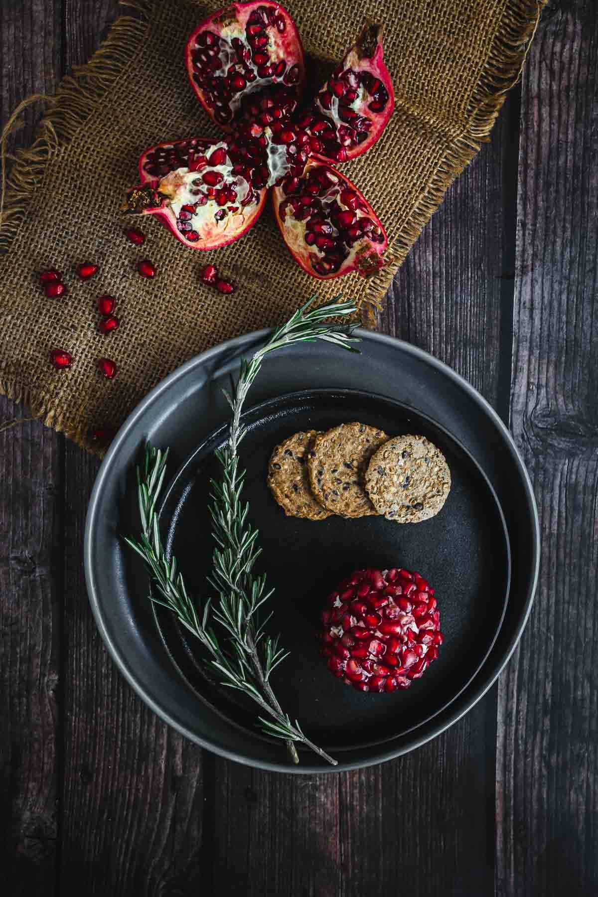 A black plate with crackers, rosemary, and a pomegranate-topped cheese ball beside a split pomegranate.